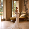 Eine Frau in einem eleganten, schulterfreien Elizabeth Passion 5817T Brautkleid mit langer Spitzenschleppe steht in einem raffinierten Raum mit hohen Fenstern, klassischem Dekor und einer alten Chaiselongue und blickt über ihre Schulter.