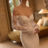 Eine Frau im Elizabeth Passion 5817T Brautkleid, einem schulterfreien, langärmeligen Spitzenhochzeitskleid, steht an einem von natürlichem Licht beleuchteten Fenster; im Hintergrund sind eine grüne Couch und eine Lampe zu sehen.