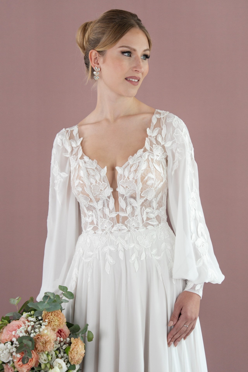 Eine Frau in Bridonista V26-A07, einem weißen, langärmeligen Brautkleid mit floralen Spitzendetails, hält einen Blumenstrauß und blickt zur Seite vor einem zartrosa Hintergrund.