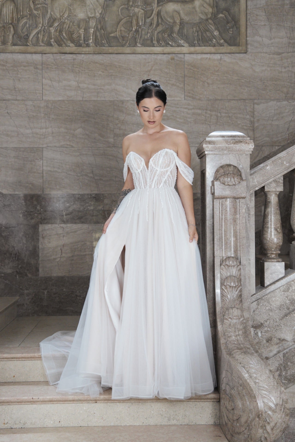 Eine Frau trägt das elegante Bridonista V26-A18: ein schulterfreies weißes Brautkleid mit transparenter Korsage und fließendem Rock. Auf Marmortreppen mit kunstvollem Steinwerk-Inspiration für moderne Brautmode.