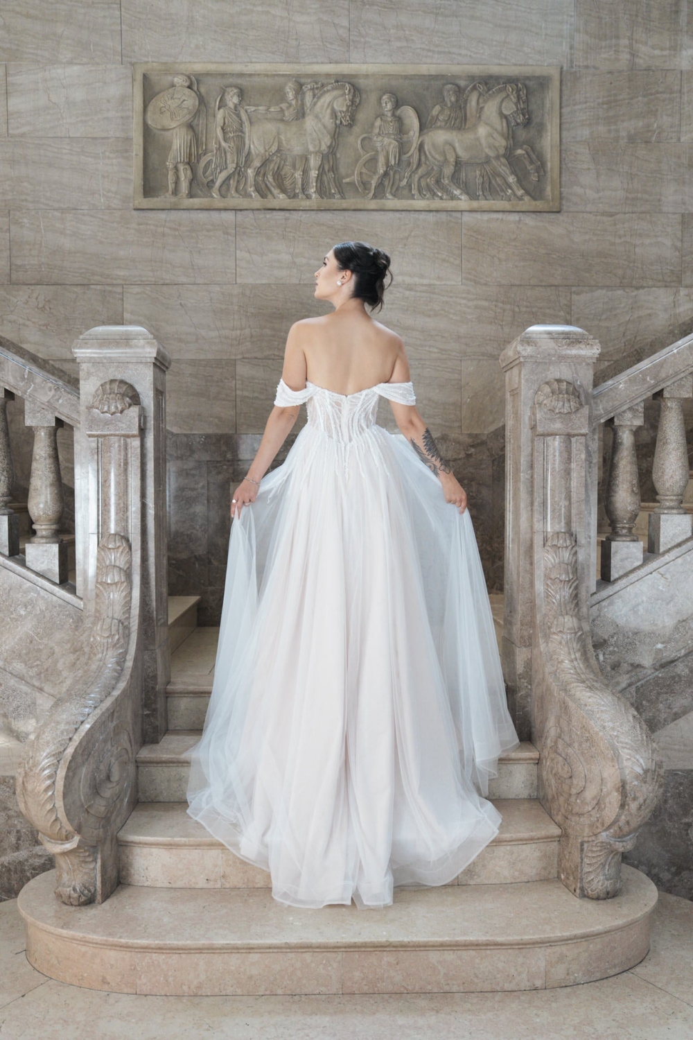 Eine Frau in einem weißen Bridonista V26-A18 Brautkleid steht auf einer Marmortreppe mit kunstvollem Geländer und wendet sich ab. Eine klassische Reliefskulptur über ihr unterstreicht die zeitlose Eleganz dieses Brautmoden-Looks.